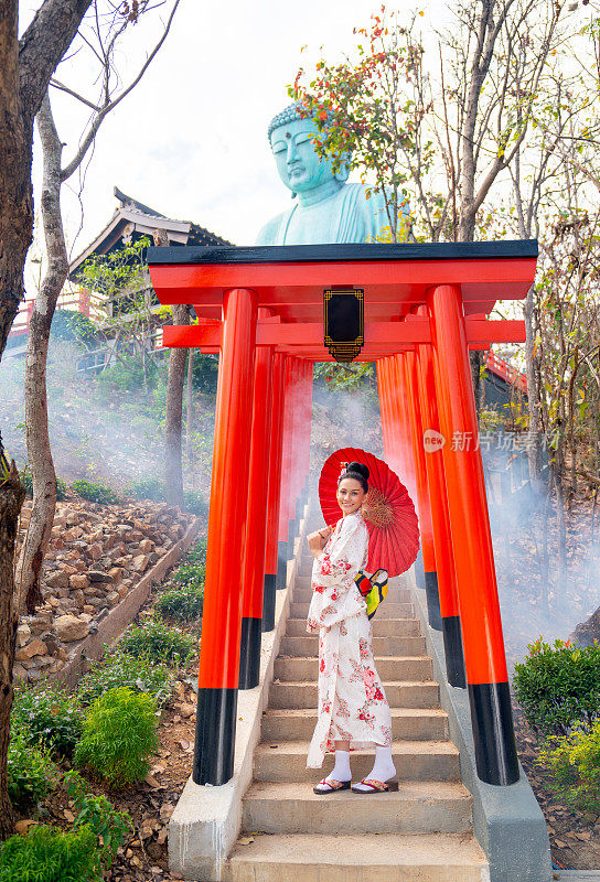 在泰国的一个旅游景点，穿着日式服装的漂亮女人手持红色雨伞站在红色拱形入口的楼梯上。