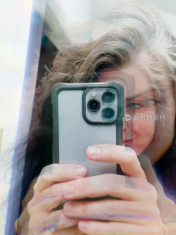 中年妇女用智能手机对着镜子拍照的抽象自拍视角。