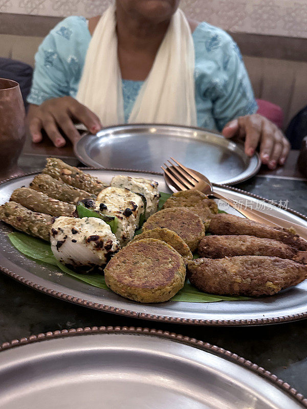 餐桌上摆放着印度小吃的金属拼盘，包括印度奶酪、炸玉米片和烤肉串，上菜的勺子和叉子，金属盘子，面面相认的印度妇女穿着蓝色的萨尔瓦·卡米兹传统服装，戴着白色的杜帕塔(围巾)。