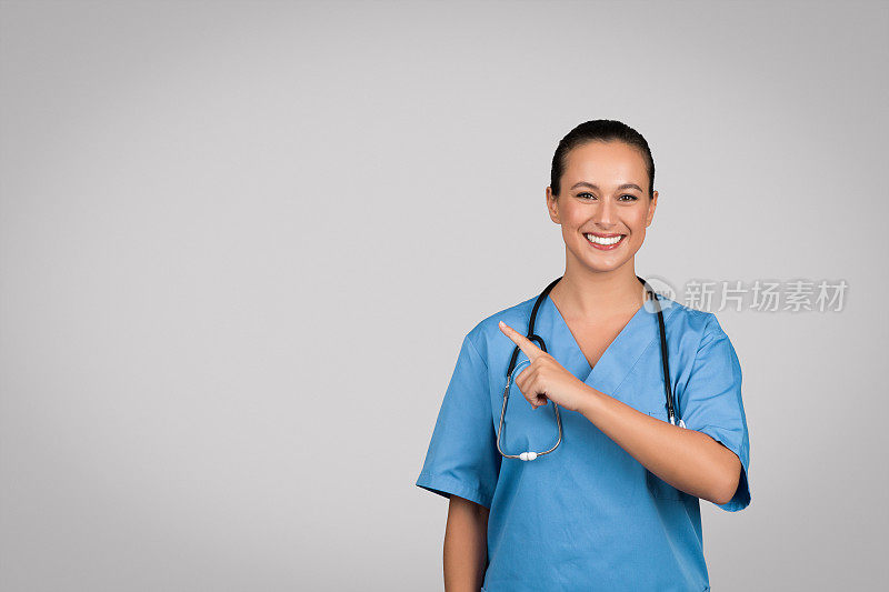 阳性白人女医生外科医生穿着蓝色制服指着灰色背景的拷贝空间
