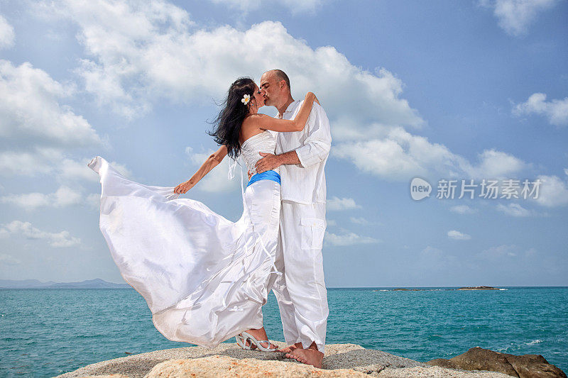 一对新婚夫妇站在海滩的一块岩石上接吻。