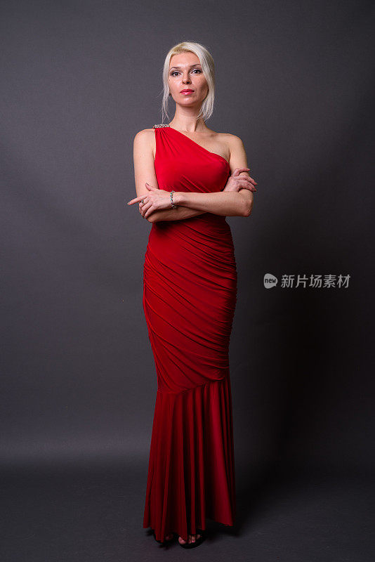 漂亮的金发女人穿着红色的不对称长裙对抗灰色的背景
