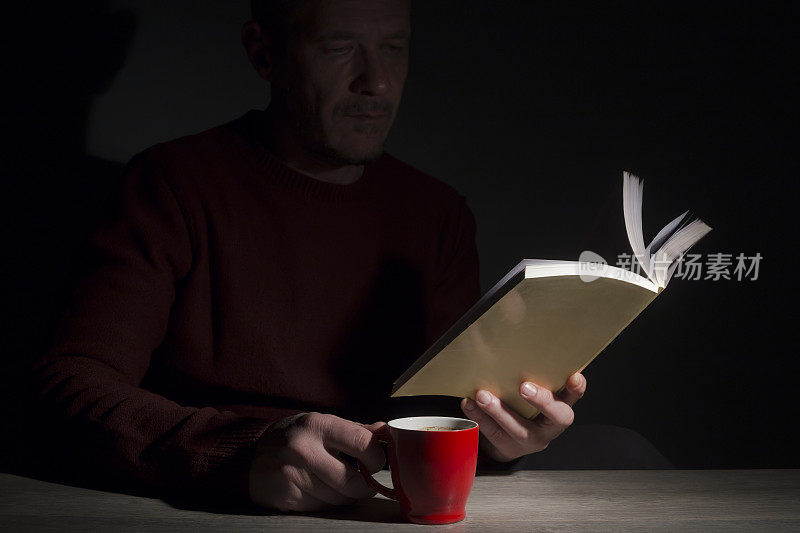晚上在昏暗的房间里，一个男人坐在桌子旁，看书，喝咖啡