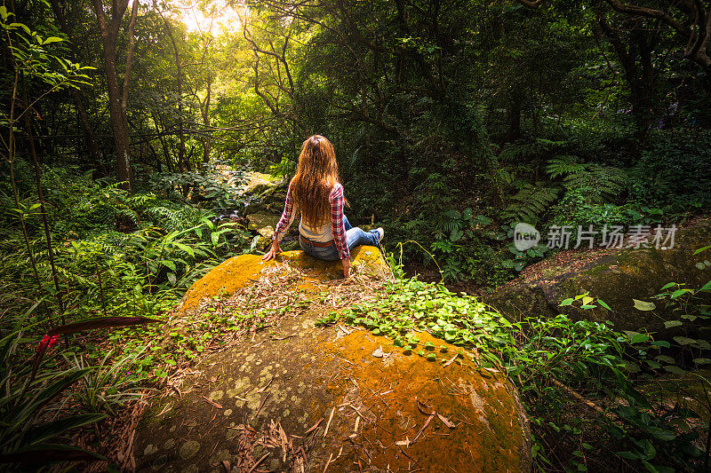 在台湾台北，一名妇女坐在长满青苔的岩石上，小溪沿着元觉瀑布徒步旅行