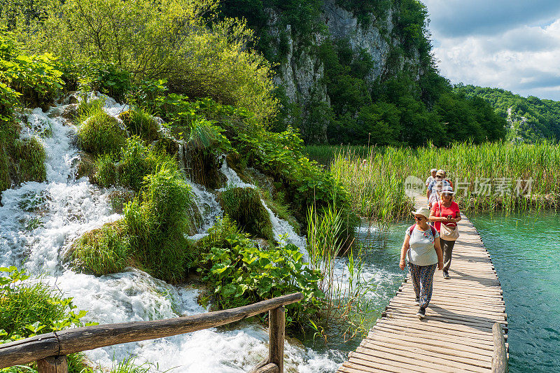 在克罗地亚的Plitvice湖国家公园，徒步旅行者经过瀑布，水流冲下进入蔚蓝色的湖泊Kaluđerovac
