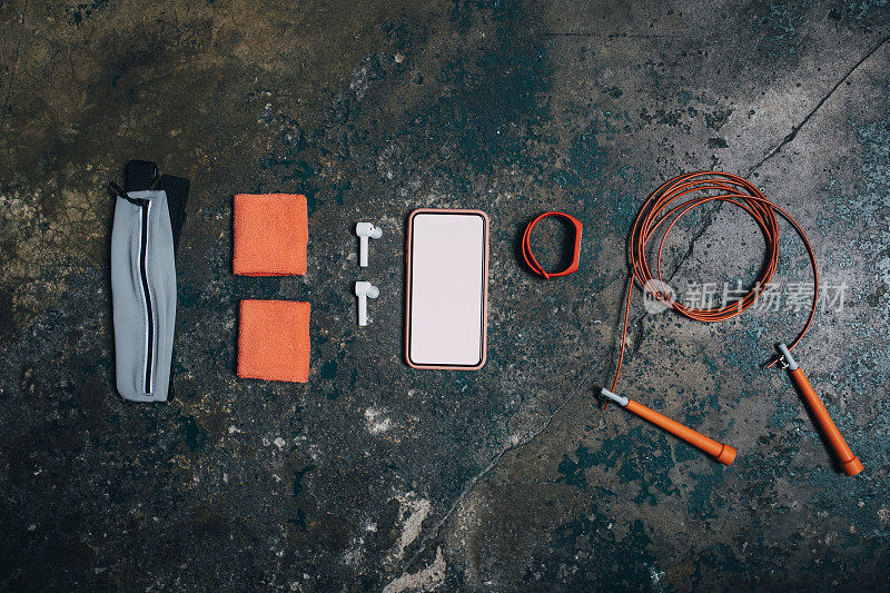橙色健身设备:一个干净的平躺在头顶上，上面有一个黑屏的智能手机，周围是一块智能手表、一根跳绳、耳机、吸汗带和一个腰带包