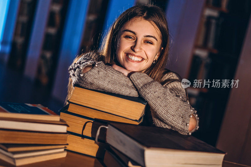 快乐微笑的女同学已经完成了考试准备工作。晚上，那女孩倚在图书馆的一堆书上
