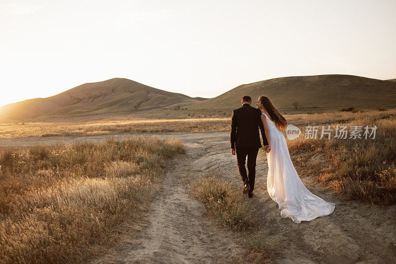新郎和新娘手牵着手走在一条小路在夕阳下的山在金色的光。