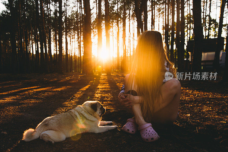 一个年轻的女人和她的狗在松树林里遇见了明亮的日落