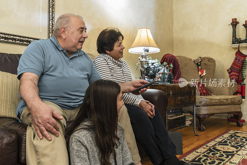 舒适的家庭。爷爷奶奶，老两口，和他们的孙女一起在为圣诞节装饰的客厅里看电视。