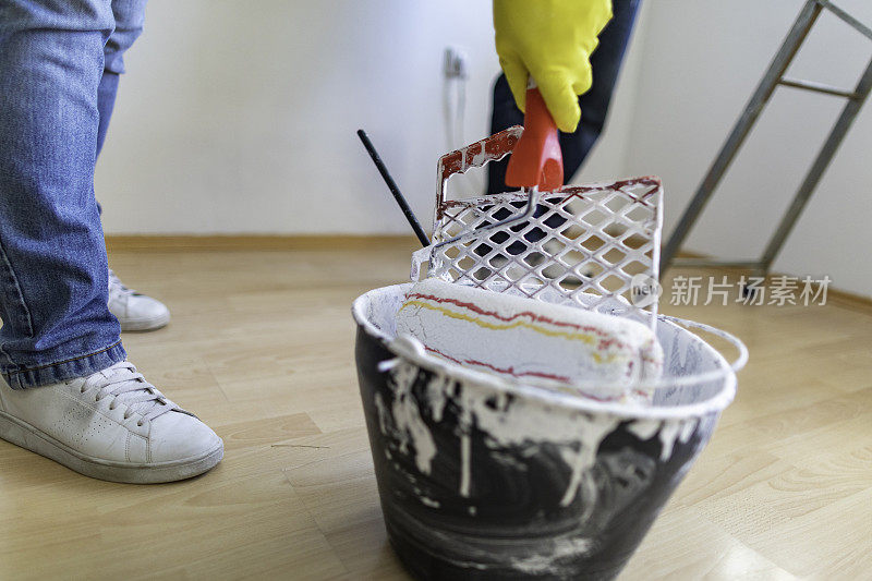 和丈夫在家里刷墙时，一位妇女在桶里蘸着油漆滚筒刷白漆