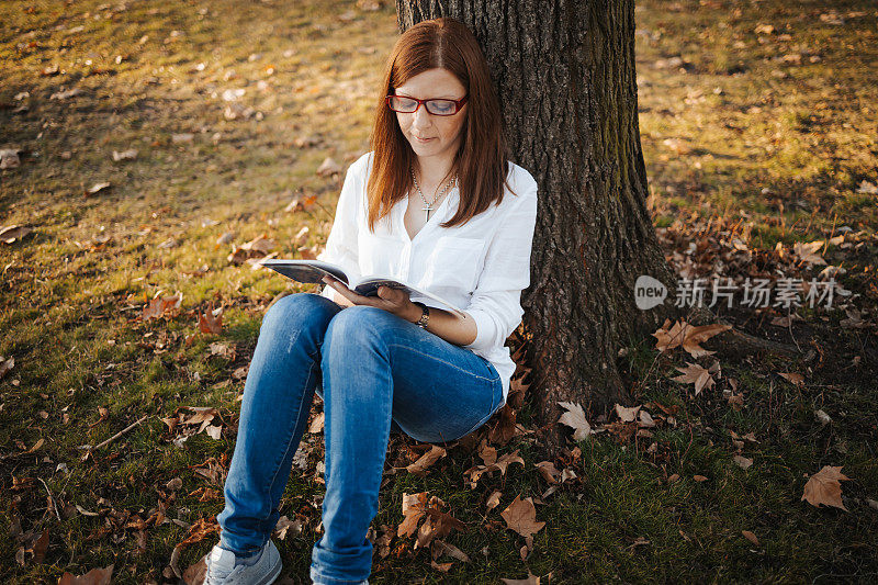 一个漂亮的女人在公园里靠在一棵树上看书