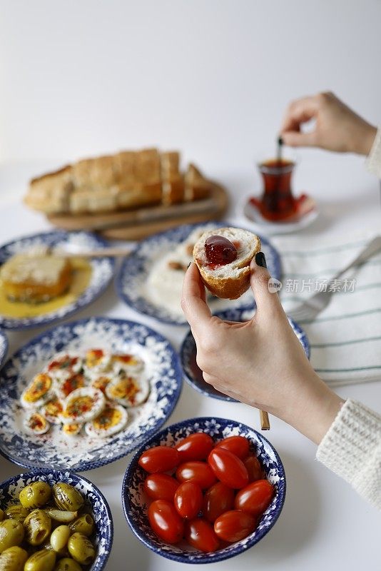 年轻的白人妇女在白色的桌子上吃着传统的土耳其早餐，一只手拿着一片面包和草莓酱，另一只手拿着土耳其红茶。早餐在家里。
