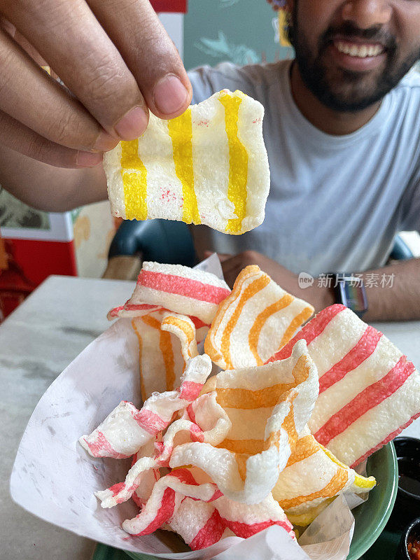 图像印度人拿着黄色条纹虾饼干，条纹，虾饼干在碗，橙色，红色和黄色条纹淀粉零食，大理石桌面背景，重点前景