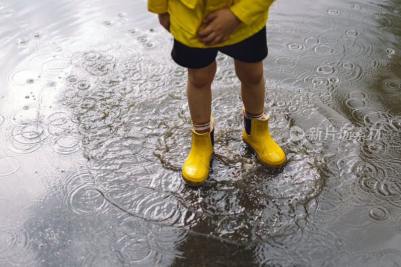 春天的一天，可爱的小男孩穿着雨衣和胶靴走过水坑