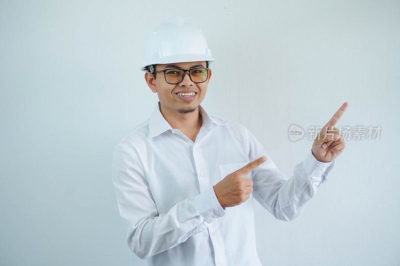 年轻的亚洲建筑师男子戴着白色安全帽，看着相机，手指指向左侧，孤立在白色背景上