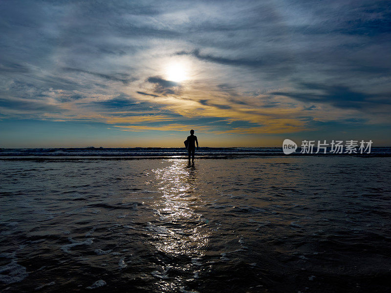 一个年轻而健壮的男孩在日落时分穿过海滩上的水去冲浪