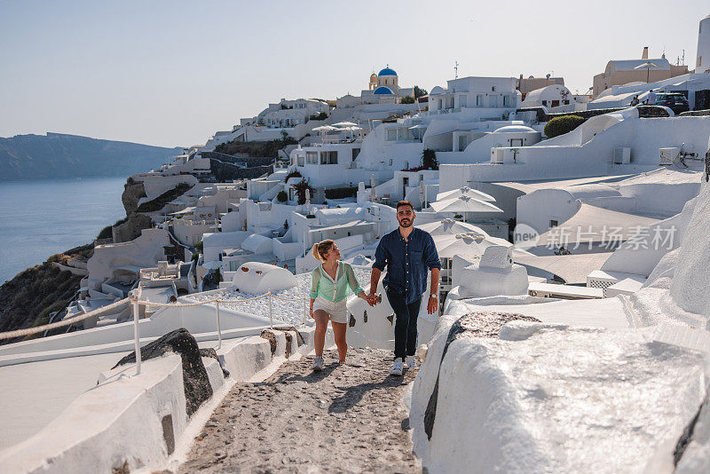 迷人的年轻白人游客夫妇手牵着手在圣托里尼岛散步