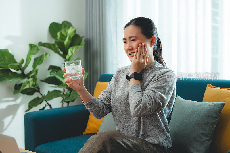 亚洲女性坐在客厅的沙发上，喝了凉水，摸了摸脸颊，就会感到牙齿敏感。