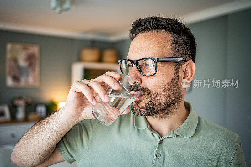 一个看起来年轻又成熟的男人站在客厅里，用杯子喝着新鲜干净的水，关心液体的摄入量