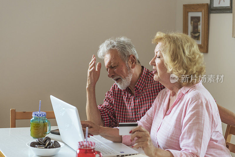 网上购物是最好的购买方式。幸福的老夫妇谈论使用笔记本电脑一起吃早餐，惊讶兴奋的妇女看着电脑屏幕上显示微笑的中年丈夫在网上购物在网站上销售