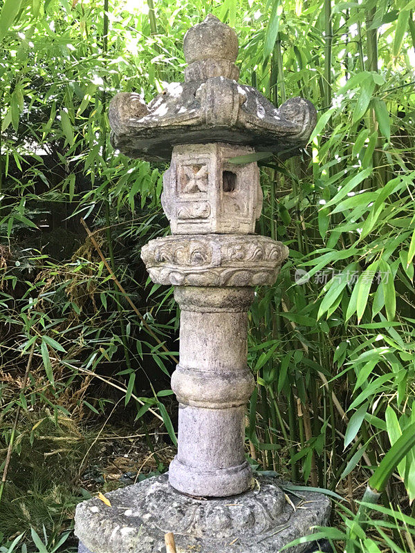 大型花岗石柱灯笼的形象在东方日本花园与观赏雕刻和柱竹背景，风化的苔藓和地衣生长表面看起来古老，禅宗灯笼独立块
