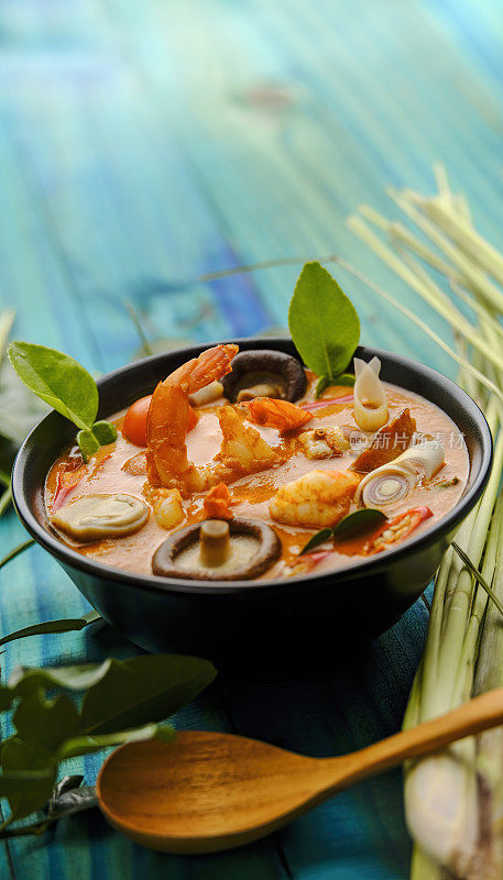 在国际上享有盛名的香辣泰式椰子汤“冬阴功”，与整个柠檬草，青柠叶和勺子一起放在绿松石色的木板桌子背景上。
