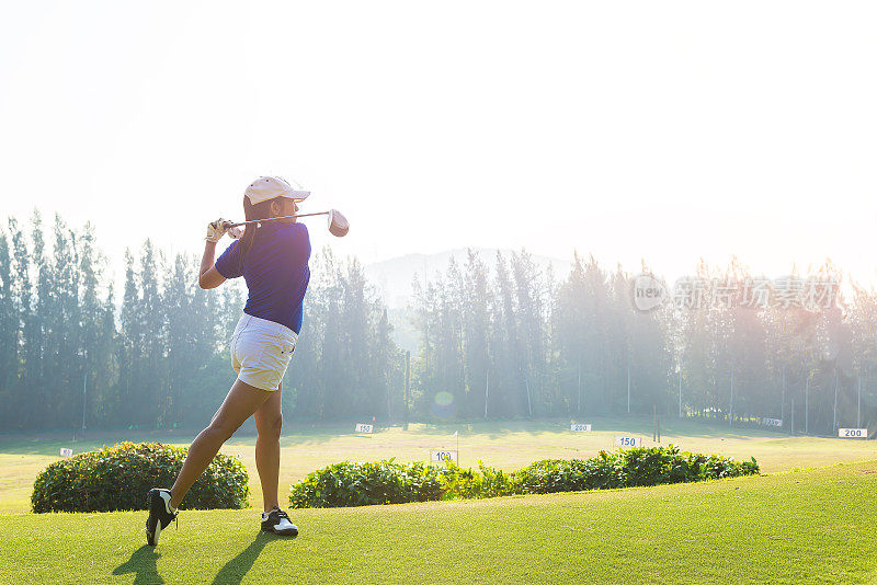 女子高尔夫球手在木杆末端挥杆的动作中，将高尔夫球从开球处击向前方的球道，夕阳的景色在背景中