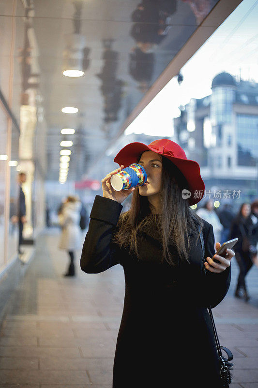 年轻女子戴着红帽子在街上一边喝着咖啡一边用智能手机发短信