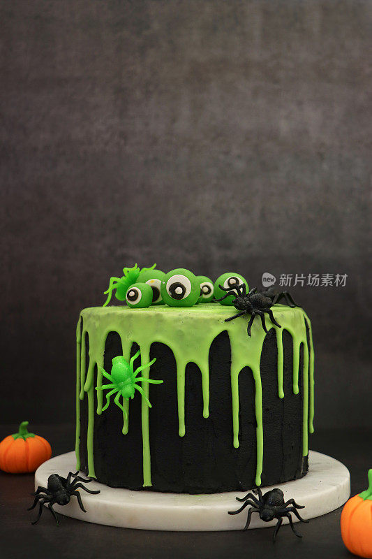 图片:自制的万圣节，分层海绵蛋糕放在大理石蛋糕上，旁边是方糖南瓜，上面覆盖着黑色的方糖糖霜，滴着绿色糖霜的设计，顶部有可食用的眼球和塑料蜘蛛，黑色背景，聚焦前景