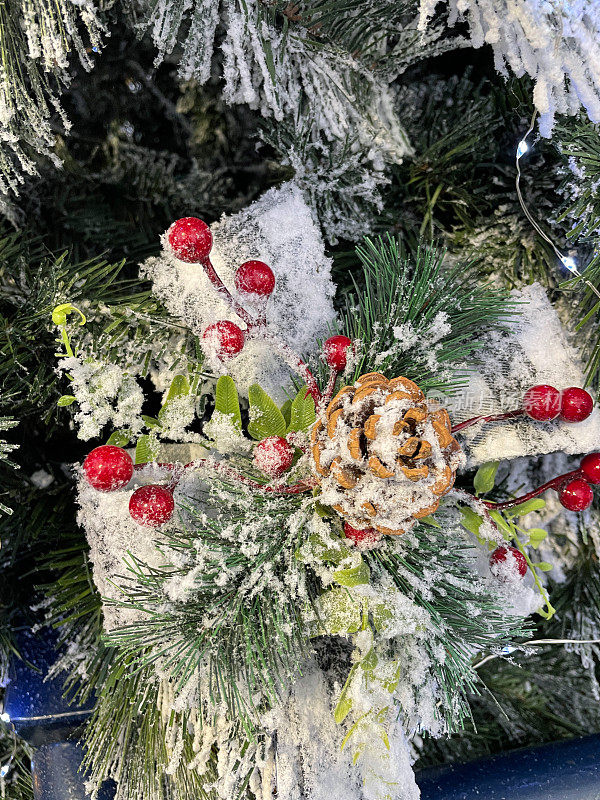 图像上白色格子蝴蝶结饰有松果和红莓饰挂在人工圣诞树的树枝上，云杉针上覆盖着人工喷雪，背景上模糊的白色神仙灯