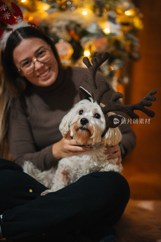 美丽的女人和她的宠物狗，摆姿势在圣诞树前鹿角发带