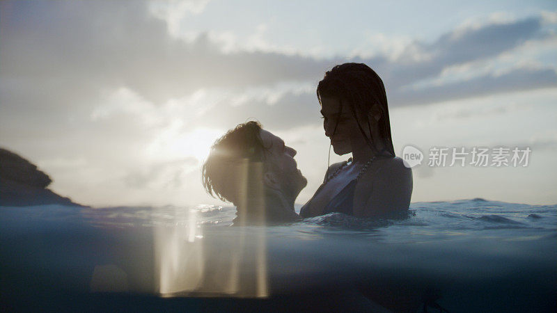 浪漫的异性情侣在美丽的金色夕阳下在海里游泳。玩耍和拥抱