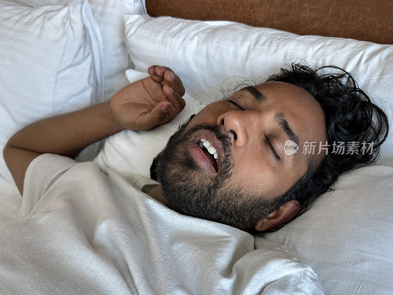 特写图像，印度男子躺在酒店房间的双人床上的枕头上，鼾声如雷，睡在白色床单下，人造革床头板，专注于前景