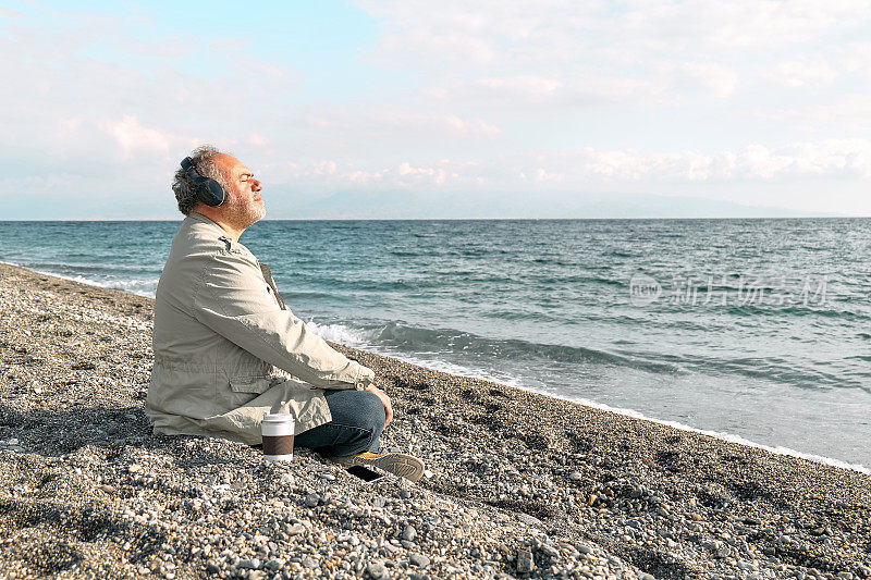 快乐的中年大胡子男子坐在鹅卵石冬季海滩上的莲花姿势，冥想和听音乐或耳机中的智能手机应用程序播客。放松的户外活动。