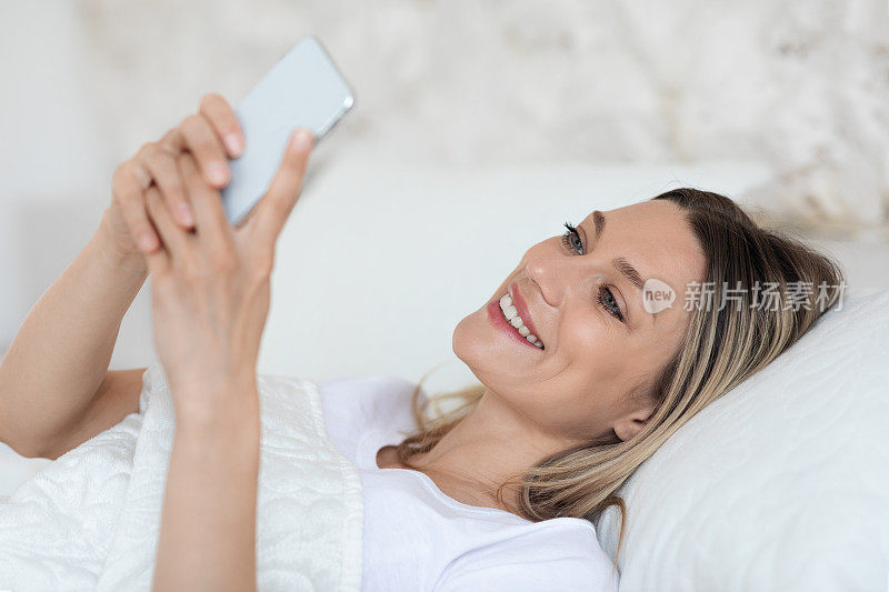 快乐的金发女人休息在床上用手机