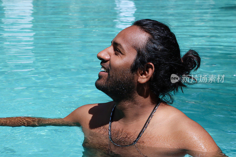 印度男子站在肩部深的清澈的水中，室外，游泳池，绿松石蓝色马赛克瓷砖被波纹水扭曲，游泳和飞溅，波涛汹涌的水面反射，重点在前景