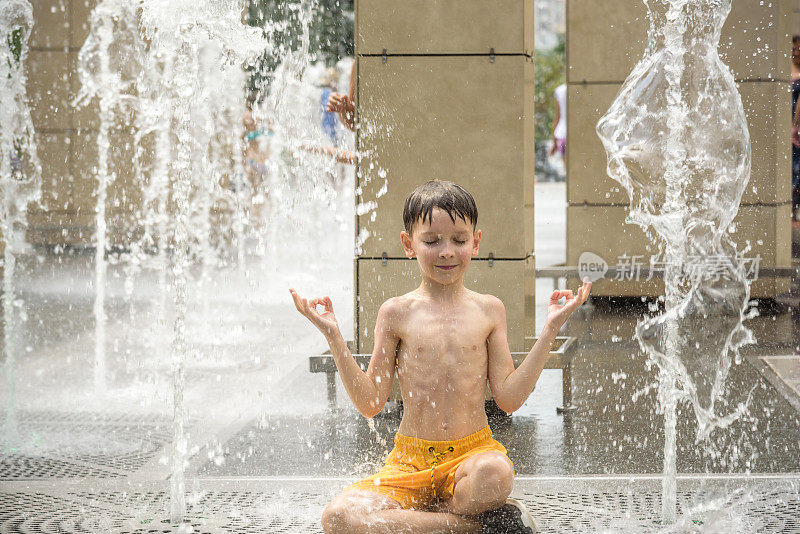 男孩在喷泉里冥想，发现禅。在炎热的夏日，一个孩子在城市喷泉边玩耍。快乐的孩子们在喷泉里玩耍。夏天的天气。积极休闲，生活方式和度假