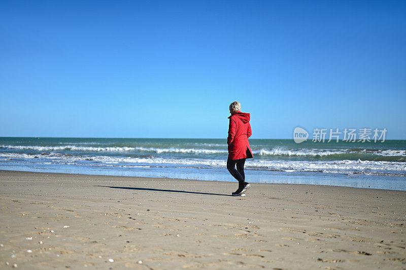 一个成熟女人在寒冷的天气里走在沙滩上的后视图。