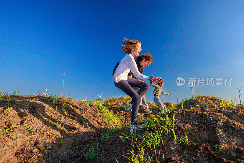 两个孩子在郊外的绿色山丘上玩耍和跳跃，旁边是风力涡轮机