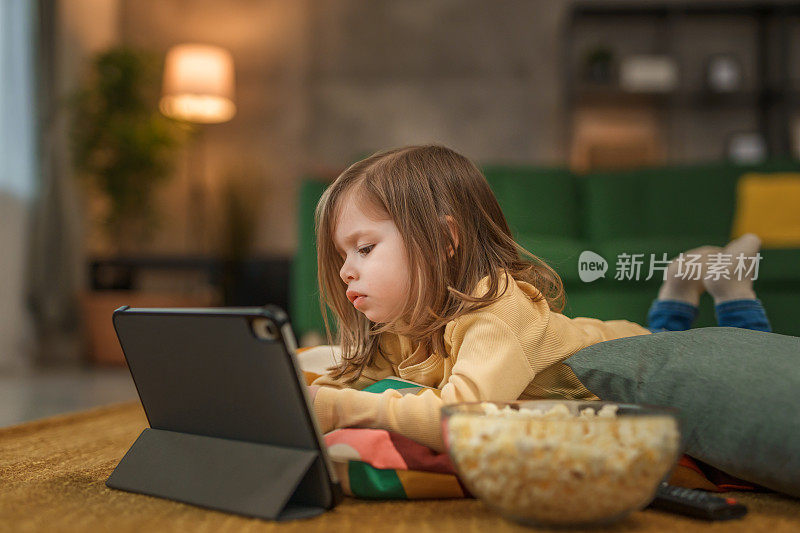 一个蹒跚学步的女孩躺在地板上用平板电脑看动画片，吃着爆米花