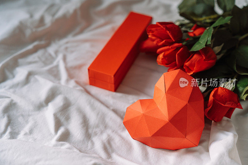 白色的床上放着纸红色的心，上面有礼物和一束玫瑰