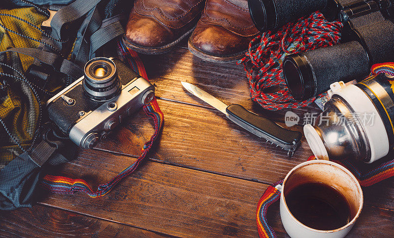 木制背景的旅行配件。旧的徒步皮靴，背包，老式胶卷相机，小刀和保温瓶