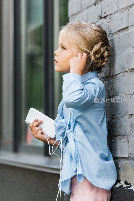 侧面的可爱的孩子戴着耳机拿着智能手机，靠在砖墙和看向别处