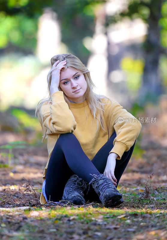美丽的金发少女盘腿坐在森林小径上，阳光斑驳