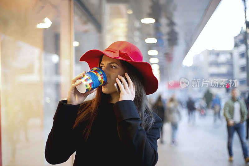 年轻女子戴着一顶红帽子在街上一边喝着咖啡一边讲着智能手机