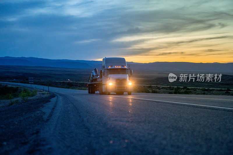 白色半卡车在四车道高速公路上超速行驶，背景是戏剧性的日落，车头灯亮着