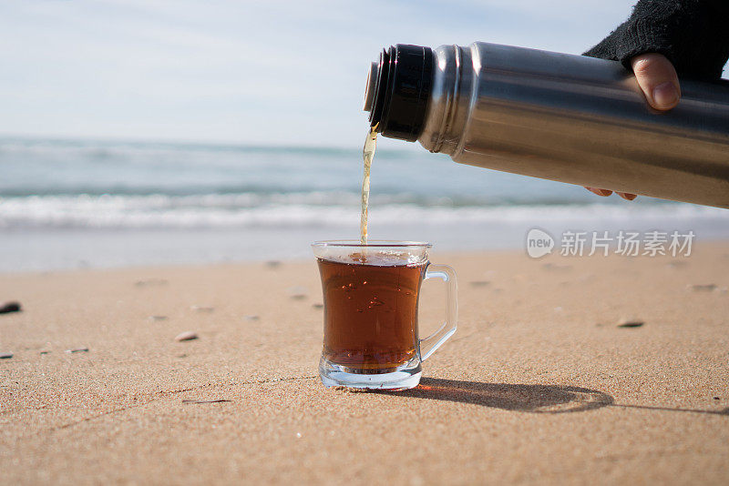 冬天，在海滩上，一名妇女正在把热水瓶里的茶倒进马克杯里。