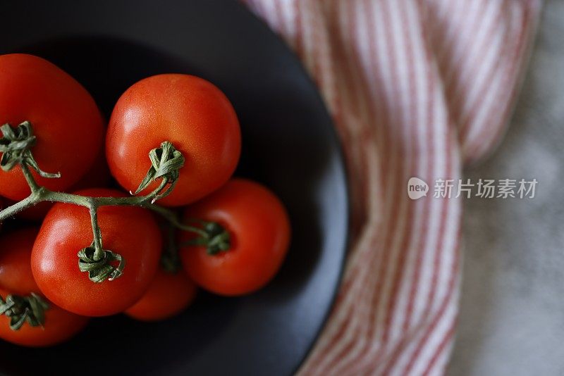 一个大的黑色陶瓷盘子的横截面从一束新鲜的樱桃番茄在一个灰色的混凝土背景，一个红白条纹布餐巾下的盘子，特写