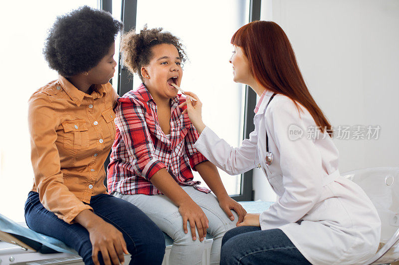 妈妈和女儿在医生办公室。白种人医生儿科医生，检查咽喉可爱的非洲少女，使用压舌板。说啊，喉咙痛，喉科概念。